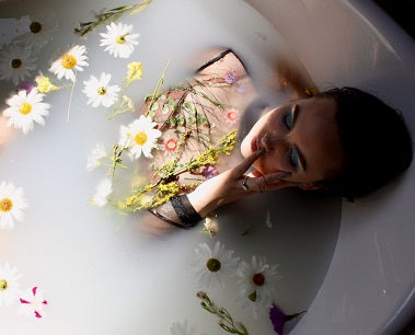 A lady relaxing in a bath of milky coloured liquid with white flowers and sprigs of herbs.